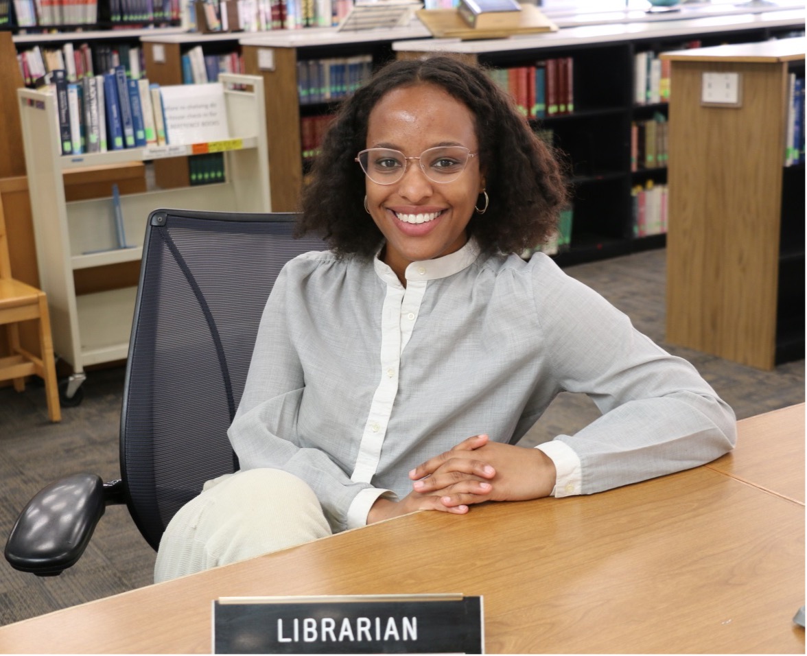 Smiling librarian sitting at library reference desk with a Librarian nameplate in foreground